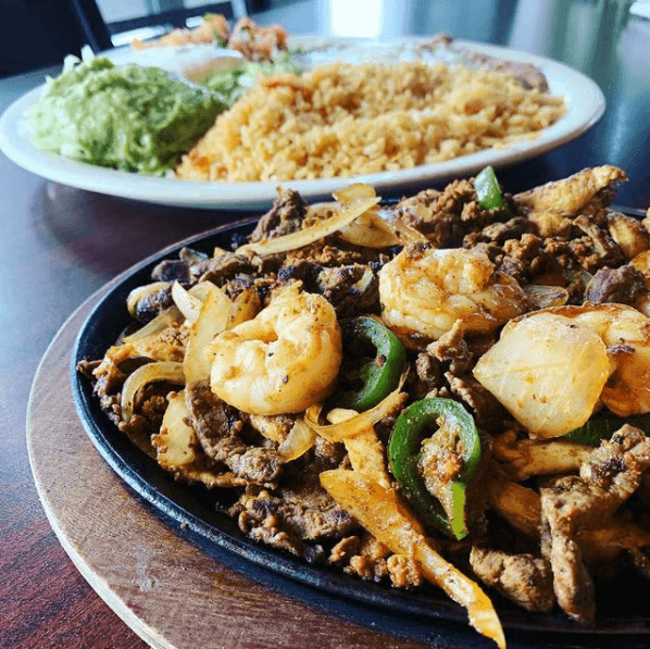 Sizzling steak and shrimp fajitas with peppers and onions on a cast-iron skillet, with rice and guacamole behind
